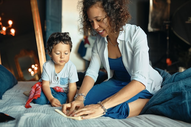 Warm cozy scene of young hispanic woman sitting on bed with her adorable son, folding paper, teaching him how to make origami.