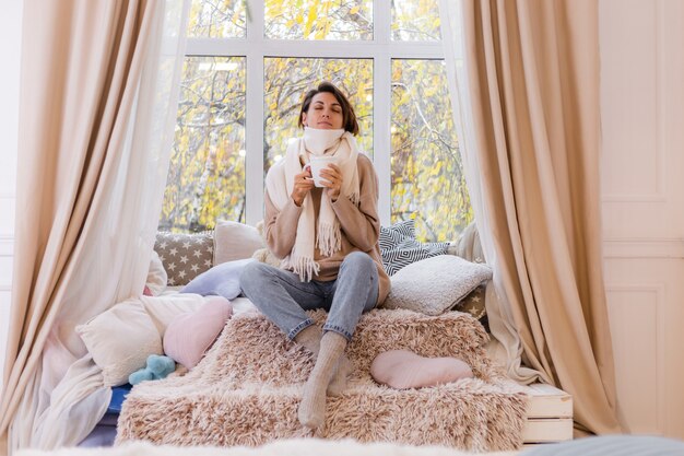 Warm portrait of woman sitting on windowsill with cup of hot tea coffee wearing sweater and white scarf