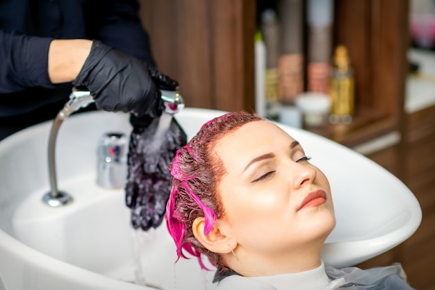 Мытье окрашенных женских волос. профессиональный парикмахер смывает с клиента краску розового цвета. молодая кавказская женщина, мыть волосы в салоне красоты.