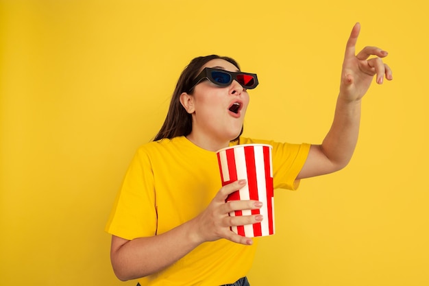 Watching cinema in 3D-eyewear with popcorn. Caucasian woman on yellow studio background. Beautiful brunette model in casual style. Concept of human emotions, facial expression, sales, ad, copyspace.