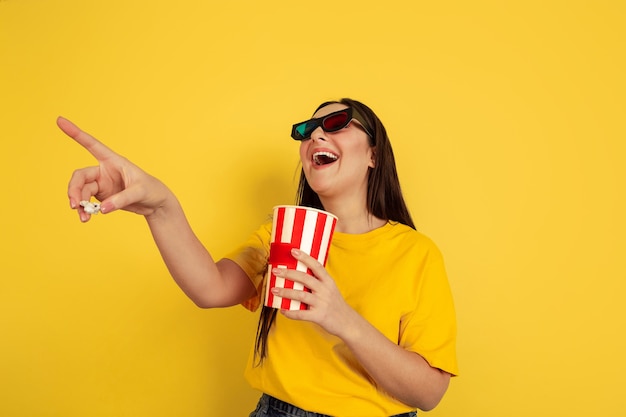 Watching cinema in 3D-eyewear with popcorn. Caucasian woman on yellow  wall. Beautiful brunette model in casual style. Concept of human emotions, facial expression, sales, ad, copyspace.