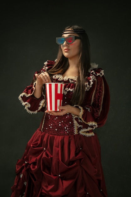 Watching cinema. Portrait of medieval young woman in red vintage clothing standing on dark background. Female model as a duchess, royal person. Concept of comparison of eras, modern, fashion, beauty.