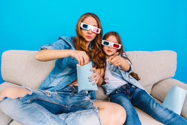 Watching movie in 3D glasses of happy mother and her daughter in jeans clothes on couch isolated on blue background. Happy family time together, eating popcorn, expressing positivity