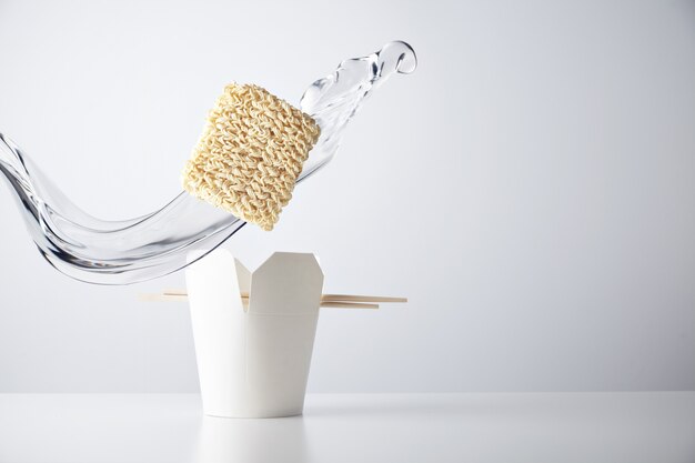 Water splash flies across the brick of dry noodles falls down inside blank takeaway box with chopsticks isolated on white commercial retail set 