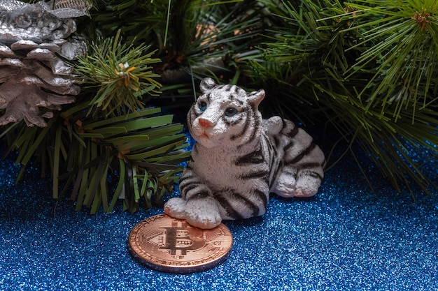 The water tiger is the symbol of the chinese new year 2022. a figurine of a lying tiger holding a bitcoin coin on a blue shiny background like on water with branches of an artificial spruce and cones.