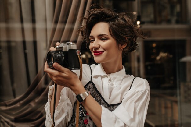 Wavy-haired woman with red lipa in light blouse holding camera in cafe. Stylish woman with brunette hair making photo inside.