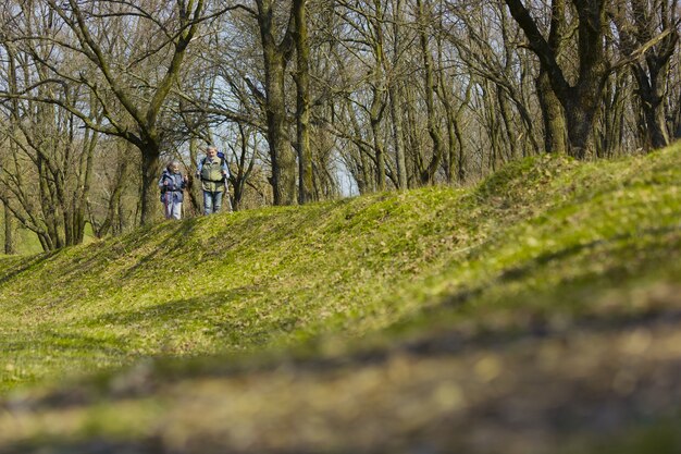 On the way together. Aged family couple of man and woman in tourist outfit walking at green lawn near by trees in sunny day. Concept of tourism, healthy lifestyle, relaxation and togetherness.