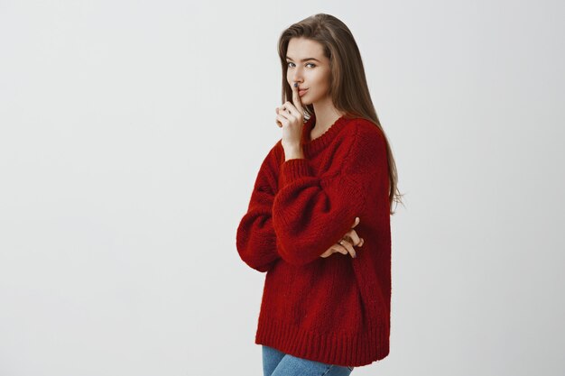 We all have secrets in closets. Portrait of romantic sensual european woman in loose red sweater, standing in profile , saying shh or shush gesture, hiding something over gray wall