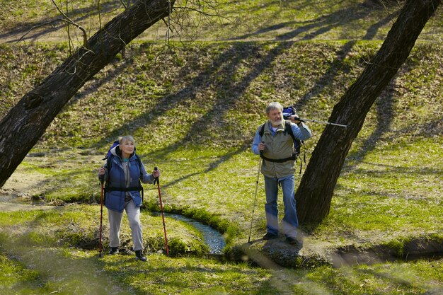 We can do it together. Aged family couple of man and woman in tourist outfit walking at green lawn in sunny day near by creek. Concept of tourism, healthy lifestyle, relaxation and togetherness.