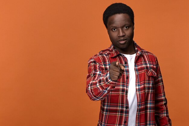 We choose you. Stylish attractive young Afro American man in plaid shirt having serious confident facial expression, pointing index finger  posing against blank orange  wall