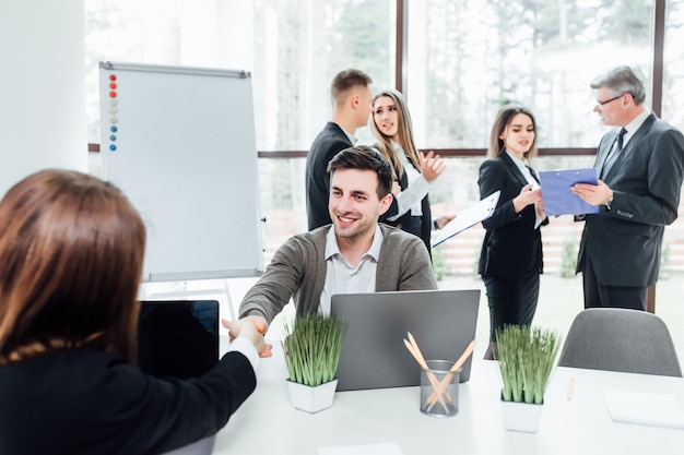 We have a deal! Men shaking hands  woman and looking at each other with smile while sitting at the business meeting with their coworkers in modern office.