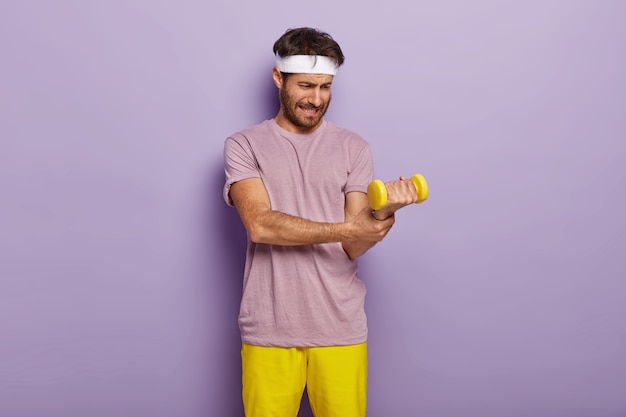 Бесплатное фото Слабый мужчина пытается поднимать тяжелые гантели, хочет быть сильным и подтянутым, регулярно делает упражнения, одет в футболку и желтые шорты.
