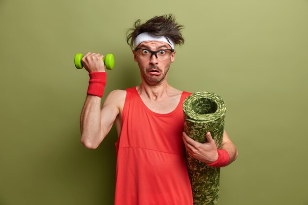 Weak man wants to become bodybuilder, raises arms with dumbbell, shocked how heavy it is, holds karemat under armpit, dressed in red sportswear, isolated on green wall. Healthy lifestyle