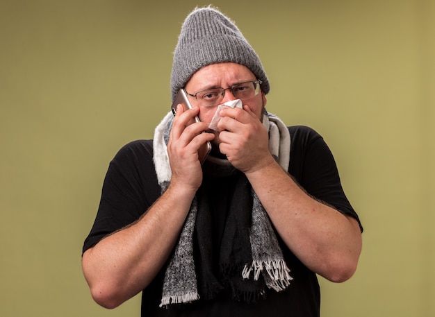 Debole maschio malato di mezza età che indossa cappello invernale e sciarpa parla al telefono asciugandosi il naso con un tovagliolo isolato sulla parete verde oliva
