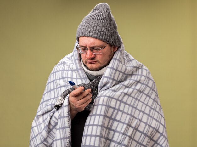 Weak middle-aged ill male wearing winter hat and scarf wrapped in plaid holding and looking at thermometer isolated on olive green wall