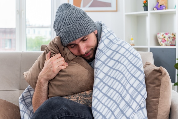 Weak and pleased young ill man wearing scarf and winter hat sitting on sofa in living room wrapped in blanket hugging pillow resting head on it with closed eyes