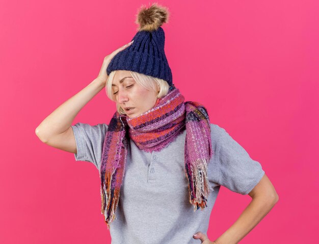 Weak young blonde ill slavic woman wearing winter hat and scarf puts hand on head