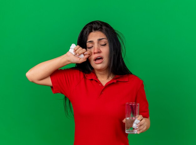 Weak young caucasian ill girl holding pack of tablets glass of water and napkin touching face with closed eyes isolated on green background with copy space