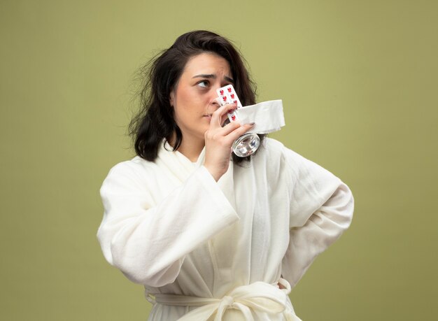 Weak young caucasian ill girl wearing robe holding pack of medical pills glass of water and napkin drinking water keeping hand on waist looking at side isolated on olive green background