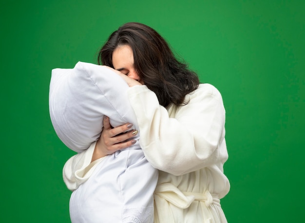 Weak young caucasian ill girl wearing robe standing in profile view hugging pillow putting head on it with closed eyes isolated on green background with copy space