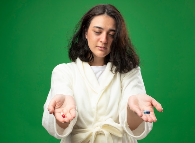 Weak young caucasian ill girl wearing robe stretching out medical capsules towards camera with closed eyes isolated on green background