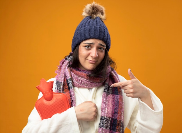 Foto gratuita debole giovane ragazza malata caucasica che indossa abito invernale cappello e sciarpa che tiene e che indica alla borsa dell'acqua calda isolata sulla parete arancione