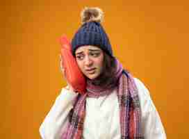 Free photo weak young caucasian ill girl wearing robe winter hat and scarf touching head with hot water bag looking at side isolated on orange background with copy space