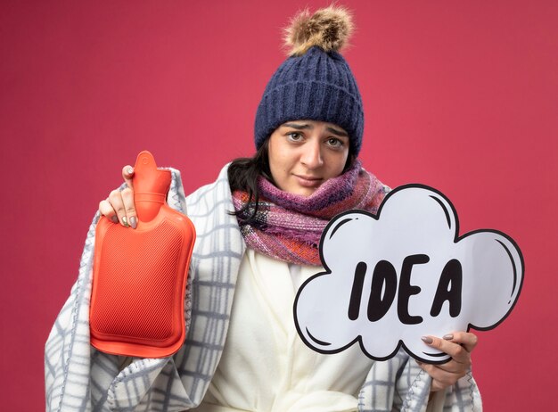 Weak young caucasian ill girl wearing robe winter hat and scarf wrapped in plaid holding hot water bag and idea bubble isolated on crimson wall