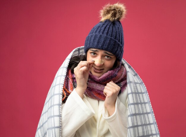 Слабая молодая кавказская больная девушка в зимней шапке и шарфе, завернутом в плед, с кулаком в воздухе, касающимся пластыря на носу, изолированном на малиновой стене