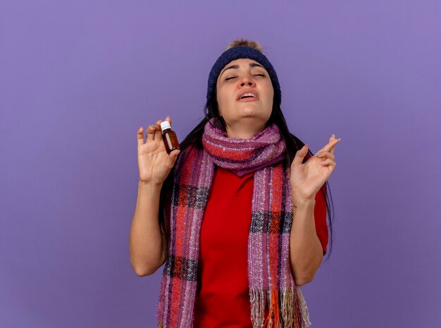 Weak young caucasian ill girl wearing winter hat and scarf holding medicament in glass doing good luck gesture with closed eyes isolated on purple background with copy space