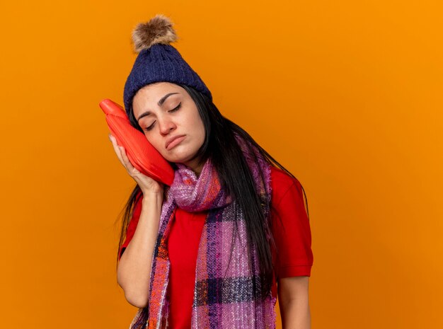 Weak young caucasian ill girl wearing winter hat and scarf touching face with hot water bag with closed eyes isolated on orange background with copy space