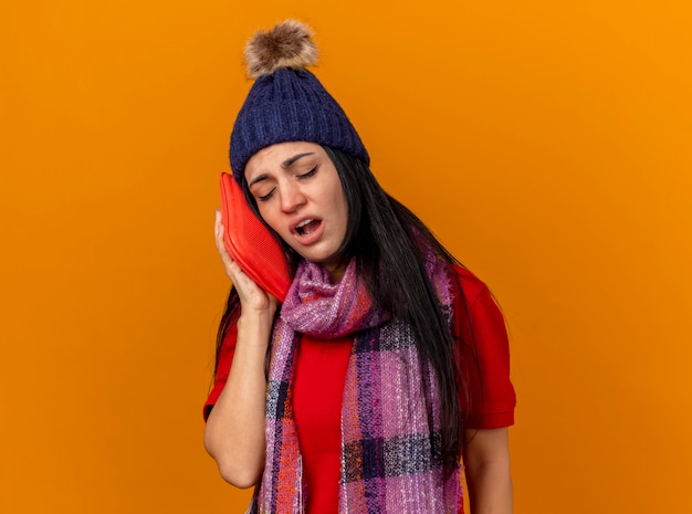 Weak young caucasian ill girl wearing winter hat and scarf touching face with hot water bag with closed eyes isolated on orange wall with copy space