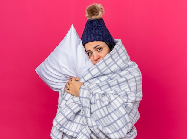 Weak young caucasian ill girl wearing winter hat and scarf wrapped in plaid hugging pillow putting head on it looking at camera isolated on crimson background with copy space