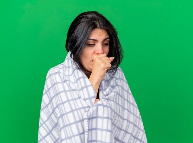 Weak young caucasian ill girl wrapped in plaid coughing keeping fist on mouth looking down isolated on green background with copy space