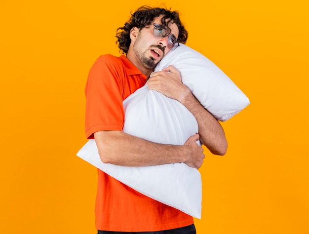 Weak young caucasian ill man wearing glasses hugging pillow looking up isolated on orange background with copy space