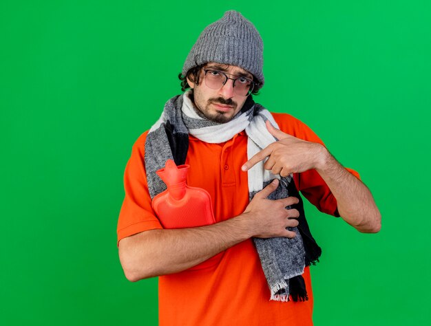 Weak young caucasian ill man wearing glasses winter hat and scarf holding and pointing at hot water bag looking at camera isolated on green background with copy space