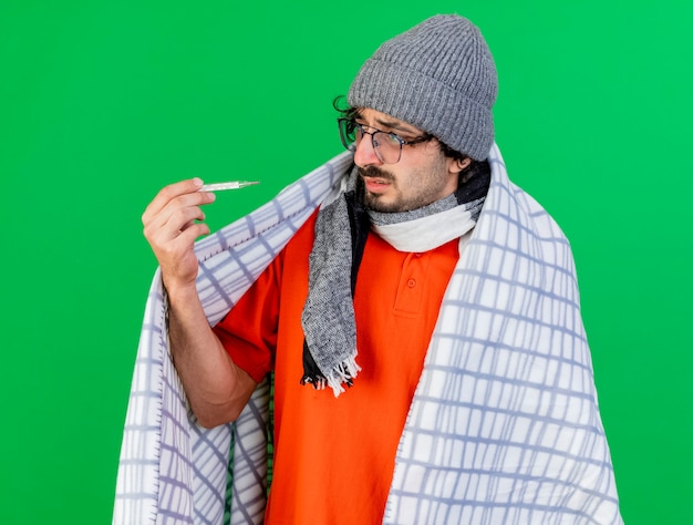 Foto gratuita debole giovane indoeuropeo uomo malato con gli occhiali inverno cappello e sciarpa avvolto in plaid holding e guardando il termometro isolato su sfondo verde