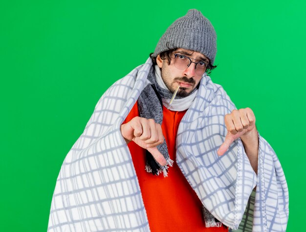 Weak young caucasian ill man wearing glasses winter hat and scarf wrapped in plaid holding thermometer in mouth  showing thumbs down isolated on green wall