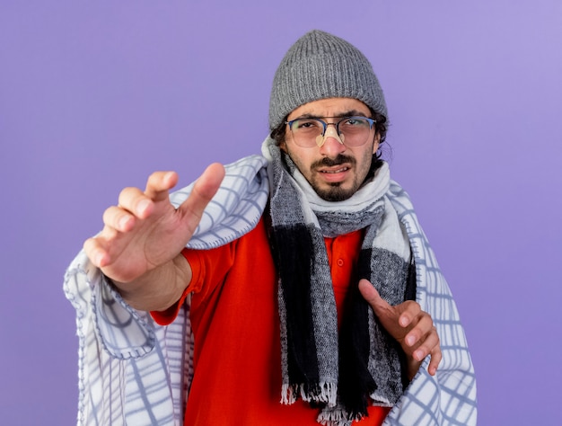 Weak young caucasian ill man wearing glasses winter hat and scarf wrapped in plaid looking at camera stretching out hands towards camera isolated on purple background