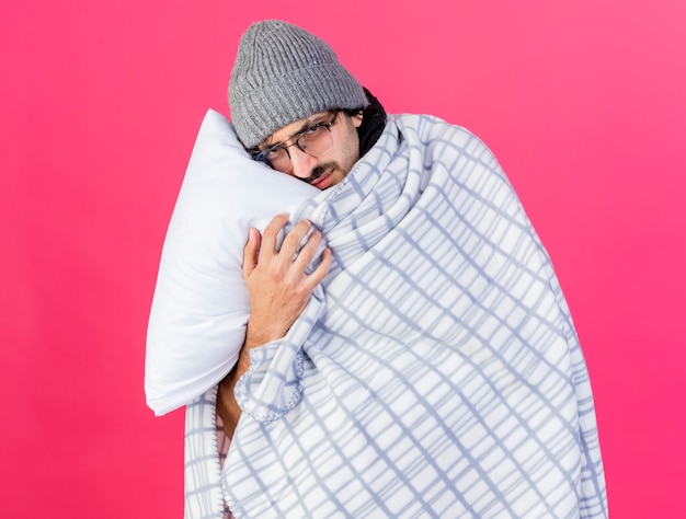 Weak young caucasian ill man wearing glasses winter hat wrapped in plaid holding pillow putting head on it looking at camera isolated on crimson background