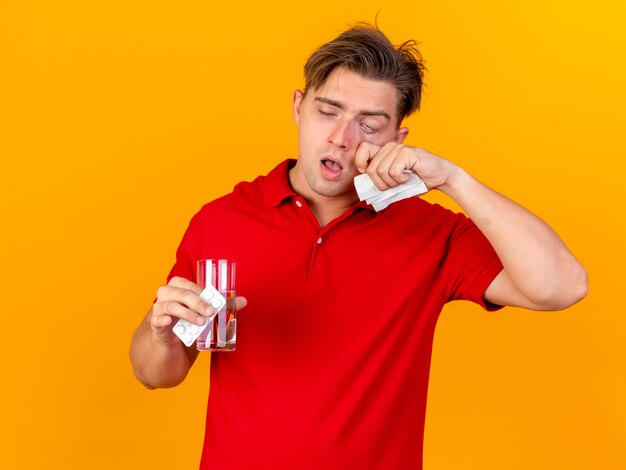 Weak young handsome blonde ill man holding pack of medical tablets and glass of water with napkin touching face isolated on orange wall