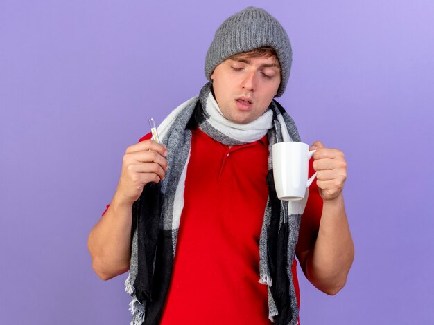 Weak young handsome blonde ill man wearing winter hat and scarf holding thermometer and cup isolated on purple background