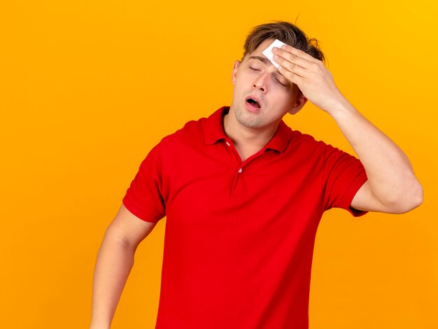 Weak young handsome blonde ill man wiping sweat with closed eyes isolated on orange background