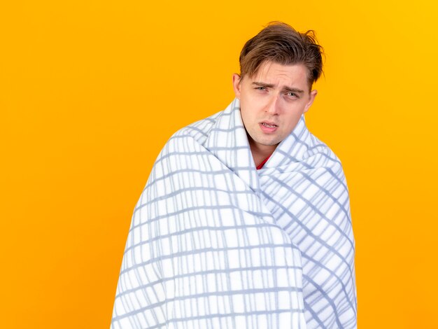 Weak young handsome blonde ill man wrapped in plaid looking at camera isolated on orange background with copy space