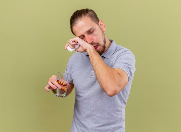 Weak young handsome slavic ill man holding pack of capsules glass of water and napkin touching face with closed eyes isolated on olive green wall with copy space