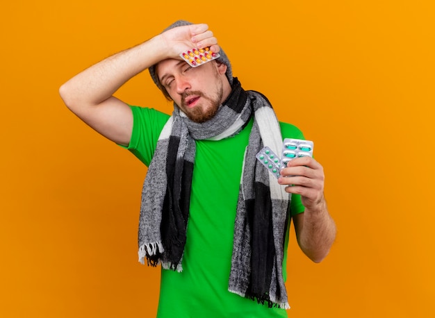 Weak young handsome slavic ill man wearing winter hat and scarf holding packs of capsules touching head with closed eyes isolated on orange wall with copy space