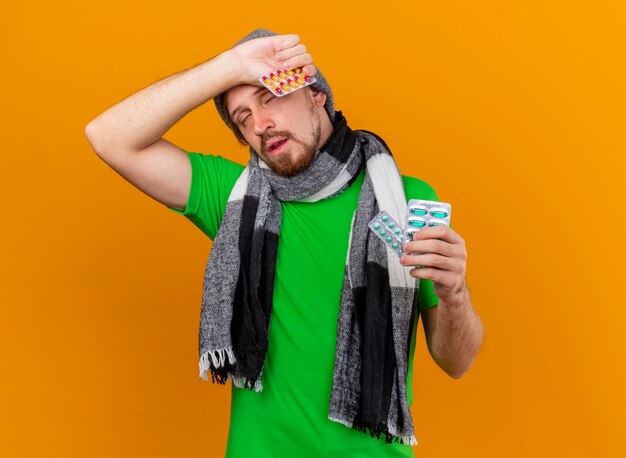 Weak young handsome slavic ill man wearing winter hat and scarf holding packs of capsules touching head with closed eyes isolated on orange wall with copy space