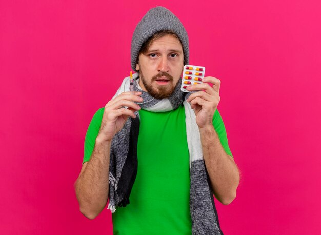 Weak young handsome slavic ill man wearing winter hat and scarf holding syringe and pack of capsules looking at front isolated on pink wall with copy space
