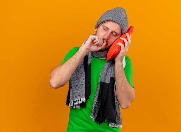 Weak young handsome slavic ill man wearing winter hat and scarf touching face with hot water bag coughing keeping fist near mouth with closed eyes isolated on orange wall with copy space