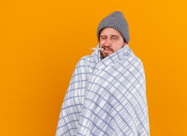 Weak young handsome slavic ill man wearing winter hat wrapped in plaid with thermometer in mouth with closed eyes isolated on orange background with copy space